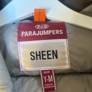 Svart dunjacka Parajumpers Sheen Y-M - Snygg svart dunjacka från Parajumpers, modell Sheen. Jackan har en glansig finish, huva och dragkedja framtill.. Perfekt för kalla dagar när du vill hålla stilen.