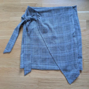 Rutig omlottkjol från Make Way - Superfin kort omlottkjol från Make Way i svart, grått och vitt rutigt mönster. Kjolen har knytband i midjan och en lätt asymmetrisk nederkant. Perfekt för dig som gillar klassisk stil med en twist.