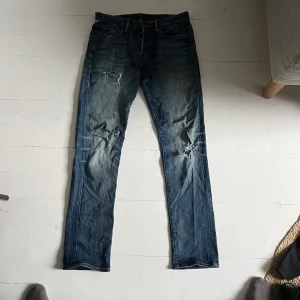 Blå slitna jeans från ralphlauren  - Säljer ett par blå jeans från ralphlauren  med snygga slitningar och hål på båda benen. Jeansen har klassisk femficksdesign, rak passform och är tillverkade i jeansmaterial. Perfekt för dig som gillar en avslappnad och cool stil.