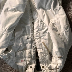 Vit parkajacka från Didriksons - Säljer en vit parkajacka från Didriksons med flera praktiska fickor och dragkedja framtill. Jackan har långärmade ärmar och detaljer som tryckknappar och broderad logga. Perfekt för kyliga dagar och har en stilren look.