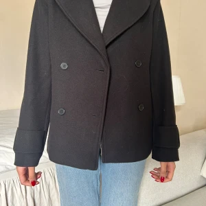 Svart skepparkavaj från Nelly - Säljer en fin svart kappa i lite tjockare material. Kort modell med dubbelknäppning och bred krage. Sitter snyggt och passar till det mesta! Knappt använd 