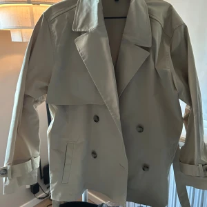 Beige trenchcoat med knappar - Snygg beige trenchcoat med dubbelknäppning, bred krage och bälte vid ärmsluten. Jackan har en loose passform och är perfekt för lager-på-lager. Klassisk och stilren design som funkar till många outfits. Har användt den 1 gång Jackan är i XL men är lien i storleken🫶🏻