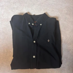 Svart skjorta från Morris Stockholm - Stilren svart skjorta från Morris Stockholm i storlek M, med klassisk krage och vita knappar. Broderad logga på bröstet och tillverkad i mjuk bomull. Perfekt för dig som gillar enkel och snygg design med diskreta detaljer.