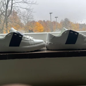 Valentino Garavani vita sneakers - Säljer ett par vita Valentino Garavani sneakers i skinn med bred mörkblå detalj över ovandelen och vita snören. Snygg och stilren design med subtila nitar runt hälen och logga i guld på insidan. Perfekta för dig som vill ha något exklusivt och trendigt.