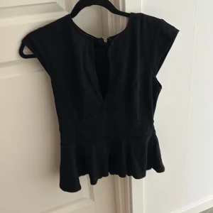 Svart peplumtop från Emamoda Paris - Säljer en svart peplumtop från Emamoda Paris i storlek L. Toppen har en snygg v-ringning, korta ärmar och en volangkant nedtill som ger en cool siluett. Perfekt att styla med jeans eller kjol för en trendig look.