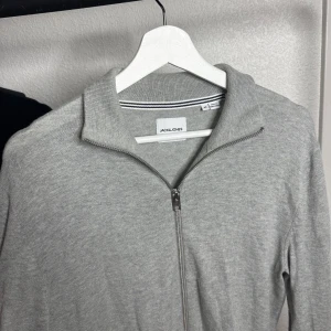 Grå kofta med dragkedja Jack & Jones - Snygg grå kofta från Jack & Jones i mjuk bomullsmix. Modellen har hel dragkedja framtill, ribbad krage och muddar samt två fickor. Perfekt för lager-på-lager och chill stil. Passar dig som gillar enkel och clean look.