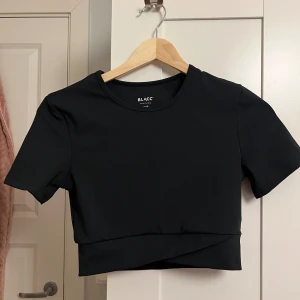 Svart croppad träningstopp från BLACC - Svart croppad träningstopp från BLACC med kort ärm och rund halsringning. Toppen har en snygg omlott-detalj nedtill och är tillverkad i stretchigt material med 80% polyester och 20% elastan. Perfekt för dig som gillar sportig och stilren look.