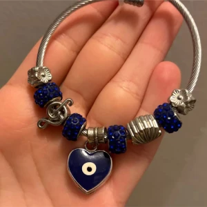 Armband med blå pärlor och hjärthänge - Unikt armband i silverfärgad metall med blå glittriga pärlor, blommor och ett hänge i form av ett blått hjärta med öga. 