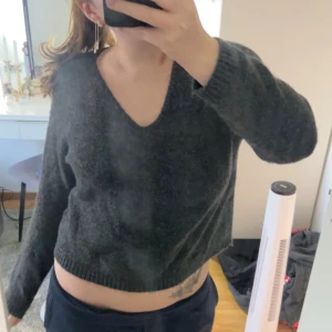 Mörkgrå stickad croppad tröja 157 - Mysig mörkgrå stickad tröja från 157 i croppad modell med v-ringad hals. Tröjan har långa ärmar och ribbade muddar vid ärmslut och nederkant. Perfekt för dig som gillar en chill och enkel stil.