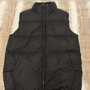 Svart dunväst från Tommy Hilfiger - Svart väst från Tommy Hilfiger med hög krage och tydlig logga broderad på insidan. Västen har en kraftig dragkedja framtill och diskret Hilfiger-flagga på bröstet. Perfekt för lager-på-lager och har två sidofickor. Använd En gång! Går att diskutera pris!