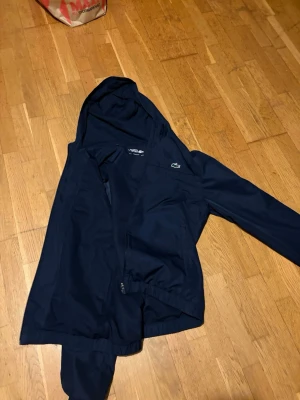 Marinblå vindjacka från Lacoste - Säljer en marinblå vindjacka från Lacoste med huva och dragkedja. Jackan har den klassiska krokodilloggan på bröstet och är tillverkad i ett lätt och smidigt syntetmaterial. Perfekt för blåsiga dagar och enkel att matcha med olika outfits. Pris kan diskuteras vid snabbaffär 