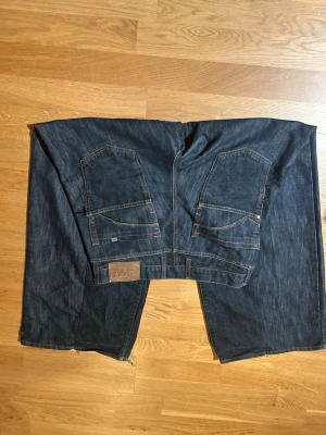 Baggy mörkblå jeans  - Omsyda brocat jeans för att få vidare ben. Mörkblå jeans med coola bakfickor. Bra skick förutom slitage vid hällen och bakfickorna. 32w i midjan och 111 cm på längden. Bara att skriva om du har några frågor :)