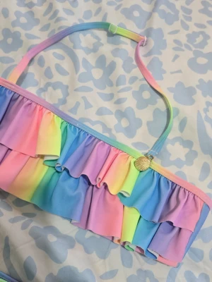 Regnbågsfärgad bikini med volanger - Säljer en bikini med regnbågsfärger i pastell och dubbla volanger på överdelen. Axelbandet är justerbart och har en liten guldfärgad snäcka som detalj. Materialet är mjukt och stretchigt syntet, perfekt för strandhäng. Storleken passar bra för 7 årig tjej. 