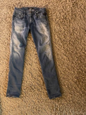 Replay Anbass jeans blå, storlek 32/34 - Säljer ett par Replay Anbass jeans i blå tvätt med snygga slitningar och klassiska fem fickor. Modellen är slim fit. Jeansen har dragkedja och knapp framtill, samt Replay-logga på linningen.