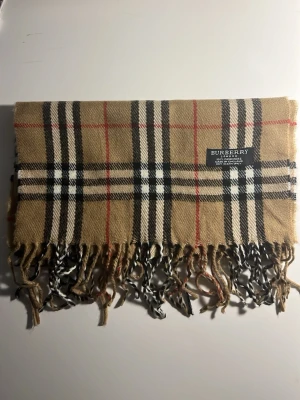 Rutig beige Burberry kashmirväst - Säljer en klassisk väst från Burberry i 100% kashmir med det ikoniska rutiga mönstret i beige, svart, vitt och röda linjer. Västen har fransar längs nederkanten och är mjuk och lyxig. Perfekt för dig som gillar tidlös design och vill ha något extra. (1A)