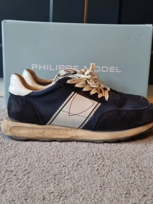 Philippe Model blå sneakers med vit detalj - Säljer ett par snygga Philippe Model sneakers i marinblått med vita och röda detaljer. Skorna har en rund tå, platt sula med unikt mönster och snörning. Ovandelen är i mocka och syntet, med vit panel och röd quiltad plös. Perfekt för dig som gillar stilrena och sportiga skor.