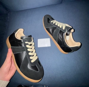 Maison Margiela svarta sneakers - Maison Margiela sneakers i svart skinn med beige detaljer och snörning. Snygg kontrast mellan den svarta ovandelen och den ljusbruna gummisulan. Klassisk rund tå och platt sula. Kommer med originalpåse och box. Perfekt för dig som gillar stilrena och lyxiga sneakers. Observa att bilderna är tagna med blixt!!
