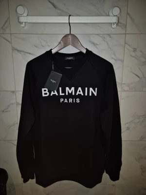 Svart sweatshirt från Balmain Paris - Svart sweatshirt från Balmain Paris med vit logotyp på bröstet. Tröjan har rund halsringning, ribbade muddar och är långärmad. Tillverkad i Italien och gjord av mjuk bomull. Perfekt för dig som gillar stilrena designer och exklusiva märken.