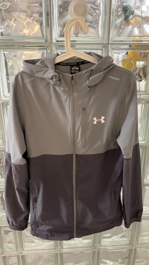 Grå vindjacka från Under Armour - Snygg tvåtonad grå vindjacka från Under Armour med huva och dragkedja framtill. Jackan har en bröstficka med dragkedja och logga på vänster sida. Perfekt för blåsiga dagar och outdoor-aktiviteter. Materialet är lätt och känns sportigt.