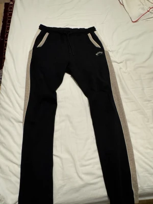 Svarta mjukisbyxor Hugo Boss - Svarta mjukisbyxor från Hugo Boss med beige sidopaneler och fickor. Byxorna har snörning i midjan och en bakficka med beige detalj. Boss-logga på vänster framsida. Perfekta för chill eller träning.