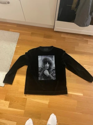 Svart sammetströja Limitato M - Svart långärmad tröja från Limitato i sammet med en stor svartvit fotoprint på bröstet. Tröjan har en mjuk känsla och är tillverkad i bomull och polyester. Perfekt för dig som vill ha något unikt och stilrent.