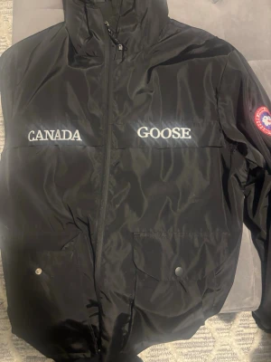 Svart vindjacka från Canada Goose - Svart vindjacka från Canada Goose med broderad logga på bröstet och patch på ärmen. Jackan har dragkedja framtill, höga fickor med lock och knapp, samt en hög krage. Perfekt för dig som vill ha en stilren och funktionell jacka.
