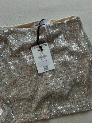 Silver paljettkjol från Bershka - Säljer en glittrig silverfärgad kortkjol från Bershka, täckt med paljetter som ger ett riktigt snyggt och festligt intryck. Kjolen har en rak passform och är fodrad med ljus insida. Perfekt för dig som vill sticka ut med lite extra bling. Silver/beige
