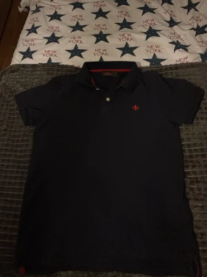 Svart piké från Morris Stockholm - Säljer en stilren svart pikétröja från Morris Stockholm i storlek M. Pikén har klassisk krage, två knappar framtill och en röd broderad logga på bröstet. Insidan av kragen har en snygg röd detalj som ger extra edge. Perfekt för dig som gillar enkel och snygg design. Passar som S