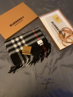 Rutig svartvit Burberry kashmirväst - Exklusiv halsduk från Burberry i klassiskt rutigt mönster med svarta, vita och gråa toner samt en röd detalj. Halsduken är tillverkad i 100% Kashmir och har fransar längs nederkanten. Perfekt för dig som vill ha en lyxig och tidlös look.