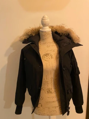 Svart Canada Goose dunjacka med päls - Svart dunjacka från Canada Goose med ikonisk patch på ärmen och fluffig päls runt huvan. Jackan har dragkedja, knappar och ribbade muddar vid ärmslut. Yttermaterialet är polyester och bomull, fodret är nylon och fyllningen består av vitt dun och coyote-päls.