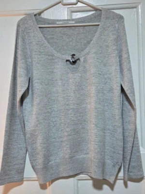 Uterqüe Premium Stickad Tröja - Ringdetalj & Båthals - Lättgrå (Strl M) - Säljer en elegant, finstickad långärmad tröja från lyxigare märket Uterqüe. Nytt skick. Tröjan har ett mjukt fall och är perfekt för chic och bekväm stil. Utmärkande detalj: Den har en smickrande båthals med en central utskärning och en dekorativ sköldpaddsmönstrad ring/spänne som håller ihop urringningen.                                    Mått: Längd från axel till nederkant är cirka 55-60 cm