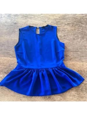 Kornblå ärmlös blus med volang - Säljer en kornblå blus med ärmlös design och rund hals. Toppen har en volang nedtill som ger en snygg peplum-effekt. Baksidan har en öppning med två knappar. Materialet är mjukt och har ett lätt glansigt utseende, perfekt för att lyfta en outfit.