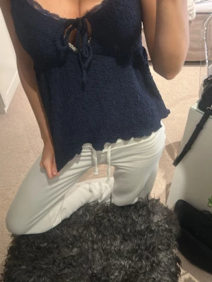 Mörkblå linnetopp med spets och knyt - Säljer en mörkblå linnetopp med smala axelband och dekorativ spetskant vid urringningen. Toppen har en snygg knytning framtill och är lätt utställd nertill. Materialet är strukturerat och känns mjukt mot huden. Perfekt för dig som gillar feminina detaljer och enkel stil.