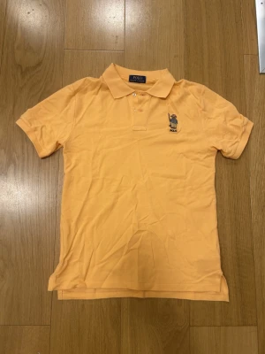 Orange pikétröja från Polo Ralph Lauren - Säljer en klassisk orange pikétröja från Polo Ralph Lauren med korta ärmar och krage. Tröjan har två knappar framtill och en broderad björn-logga på bröstet. Perfekt för en chill och stilren look. Färgen är lite mer orange lik i verkligheten. Aldrig använd.