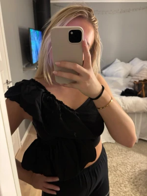 Svart oneshoulder topp från Zara - Säljer en svart oneshoulder topp från Zara med volangdetaljer och asymmetrisk design. Toppen har en axelbandslös sida och en smal axelrem, vilket ger en cool och trendig look. Perfekt att styla med höga byxor eller kjol.