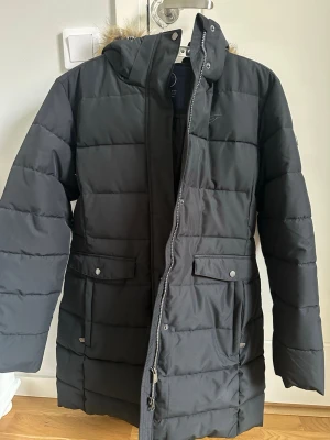 Svart jacka från Five Seasons - Svart jacka från Five Seasons med huva och fuskpäls. Jackan har flera fickor med både dragkedja och knappar, samt quiltad design. Perfekt för kalla dagar och har en stilren look som funkar till det mesta.