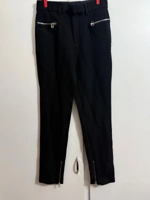 Svarta byxor med dragkedjor från Zara Woman - Svarta byxor från Zara Woman i storlek S. Modellen har smal passform med dragkedjor vid fickorna och bensluten för en edgy look. Byxorna har bälteshällor och är tillverkade i ett mjukt bomullsmaterial. Perfekt för dig som gillar detaljer och stilrena plagg.