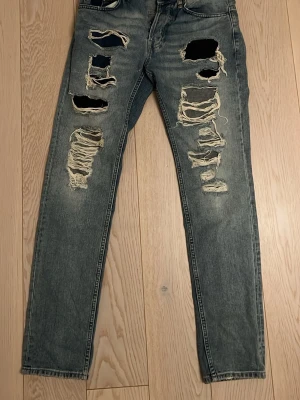 Grå slitna jeans med hål - Säljer ett par grå jeans med rejält slitna detaljer och stora hål på båda benen. Byxorna har en rak passform och är tillverkade i klassiskt jeanstyg. Perfekt för dig som gillar en edgy och avslappnad stil.