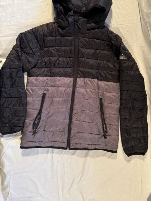Svart och grå pufferjacka McKinley - Säljer en pufferjacka från McKinley i storlek 158. Jackan är svart upptill och grå nedtill, har huva och två fickor med dragkedja. Quiltad design och hel dragkedja framtill. Perfekt för kyliga dagar och har en sportig vibe.