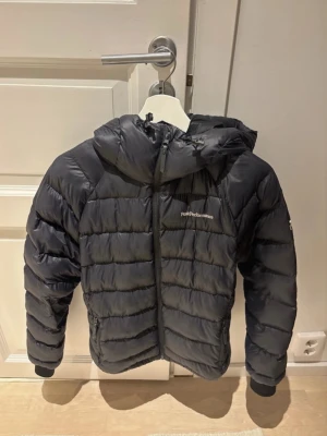 Svart dunjacka Peak Performance - Säljer en svart dunjacka från Peak Performance med huva och dragkedja framtill. Jackan har ett quiltat mönster och ribbade muddar vid ärmslut. Perfekt för kalla dagar och har en snygg, sportig look. Passar dig som gillar stilren och funktionell design.