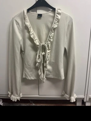 Vit topp med volang från Gina Tricot - Säljer en vit omlottopp från Gina Tricot i storlek S. Toppen har långa ärmar med volangdetaljer vid ärmslut och en djup v-ringning med volangkant. Knytning framtill ger en snygg och feminin look. Perfekt att styla med jeans eller kjol.