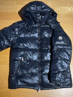 Svart Moncler pufferjacka med huva - Säljer en svart pufferjacka från Moncler med glansig finish och huva. Jackan har två snedställda fickor med dragkedja, emblem på ärmen och quiltad design. Perfekt för kalla dagar och har en snygg, modern look med Moncler-logga på insidan.  Storlek M fast sitter som en S