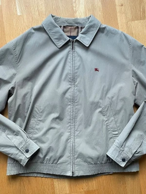 Beige herrjacka från Burberry - Snygg beige herrjacka från Burberry med klassiskt rutigt foder. Jackan har dragkedja framtill, två sidofickor och elastisk nederkant. Liten röd Burberry-logga broderad på bröstet. Perfekt för dig som gillar stilrena och tidlösa plagg.