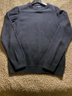 Mörkblå stickad tröja från GANT - Säljer en mörkblå stickad tröja från GANT i premium bomull. Tröjan har rund hals, ribbade muddar och ett broderat GANT-logo på bröstet. Perfekt för dig som gillar klassisk och enkel stil med snygga detaljer.