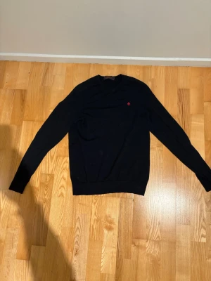 Mörkblå merinoulltröja från Morris - Mörkblå stickad tröja från Morris i 100% merinoull, med rund hals och lång ärm. Diskret röd broderad logga på bröstet ger en snygg detalj. Tröjan har en klassisk passform och ribbade muddar vid ärmslut och nederkant. Kan även vara lite sliten på vissa ställen.