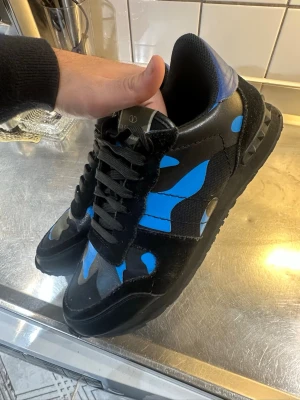 Valentino Rockrunner sneakers blå/svart - Säljer ett par Valentino Rockrunner sneakers med svart och blått camouflage-mönster. Skorna har svarta snören, rund tå och platt sula. Materialmix av skinn och textil med coola nitar på hälen. Perfekt för dig som gillar statement pieces och italiensk design.