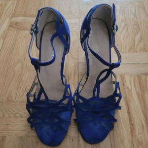 Blå klackade sandaler med strass - Säljer ett par snygga blå sandaler med hög klack och öppen tå. Skorna har ett coolt flätat mönster framtill och ett spänne med strassdetaljer vid ankeln. Materialet är mocka och sulan är platt framtill. Latin sko (salsa, bachata mm). 9cm klack. Använda bara ett par gånger. Storlek 38.