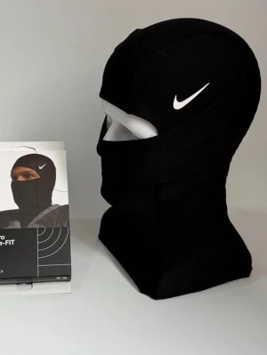 Nike Pro svart Therma-FIT balaclava - Nike Pro Therma-FIT balaclava i svart med vit Nike-logga. Tight passform som täcker hela huvudet och halsen, perfekt för kalla dagar. Tillverkad i ett syntetmaterial som andas och håller värmen. Snygg och sportig look för dig som gillar att vara aktiv utomhus.