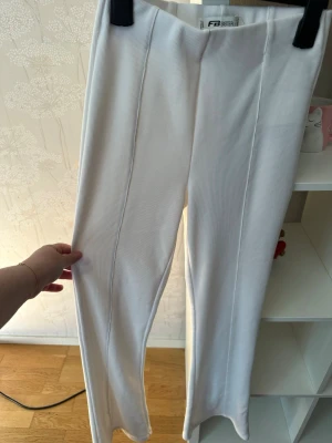 Vita ribbade byxor från FB Sister XS - Säljer ett par vita ribbade byxor från FB Sister i storlek XS. Byxorna har hög midja och raka ben med sömmar framtill som ger en snygg look. Materialet är mjukt och stretchigt, perfekt för dig som gillar bekväma och stilrena plagg.