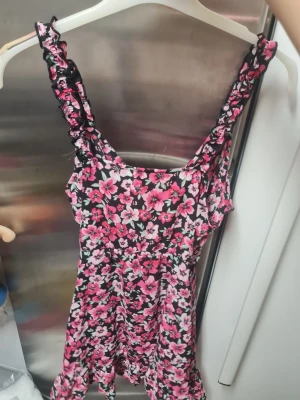 Blommig kortklänning från H&M Divided - Supersöt kortklänning från H&M Divided med svart botten och rosa blommigt mönster. Klänningen har volangdetaljer på axelbanden och en lätt utställd kjol. Perfekt för sommarens alla dagar!